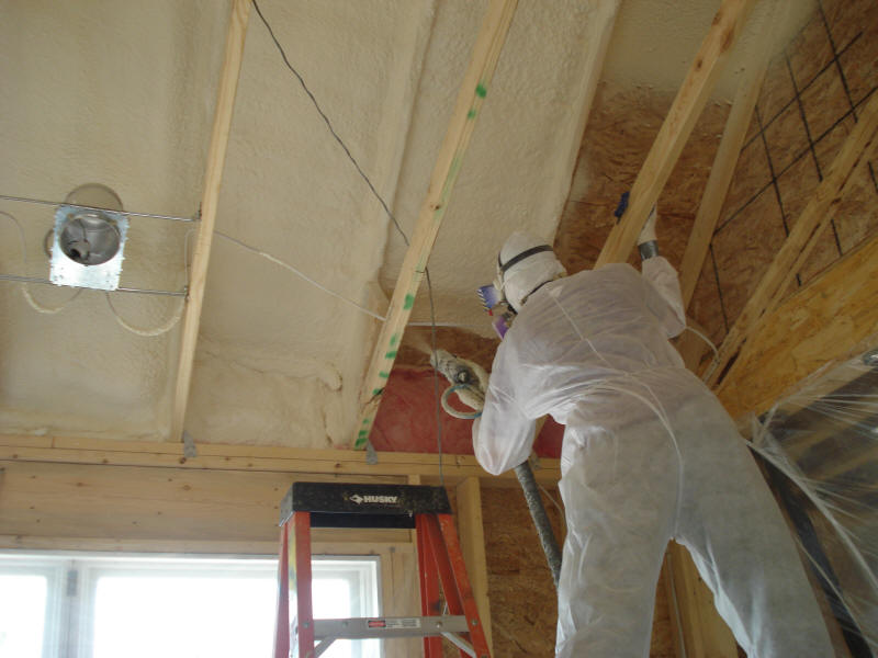 Closed cell spray foam being applied into ceiling joists by man in tyvek suit